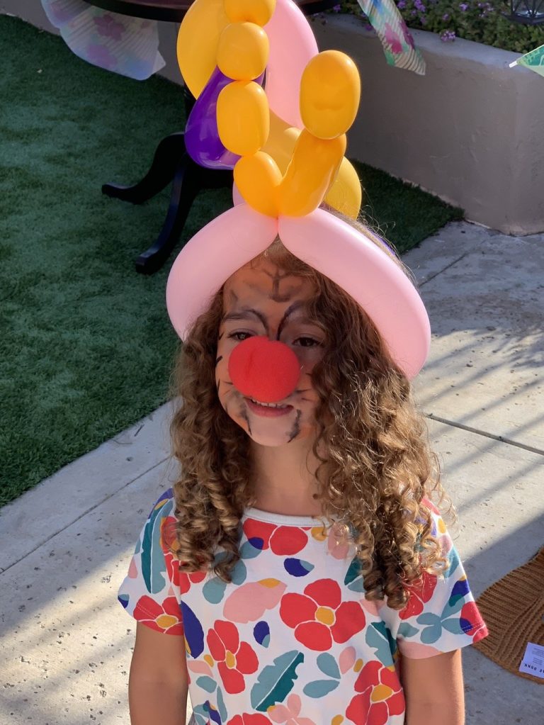 Child wearing a pink and orange balloon hat beside Sparkles