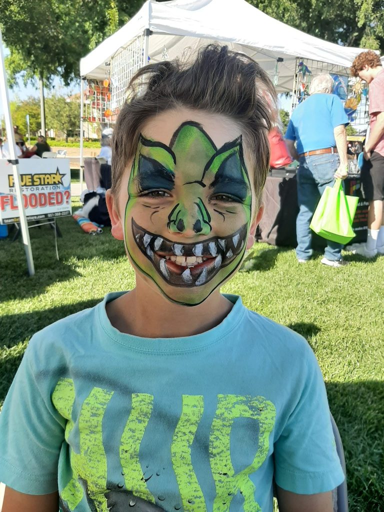 Child smiling with green dinosaur face painting at an outdoor event