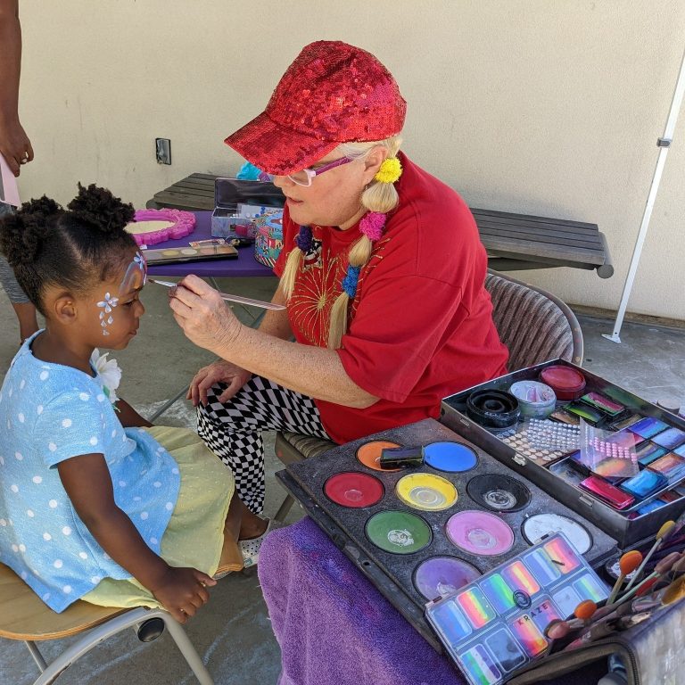 Sparkles applying face paint to a seated child at an outdoor party