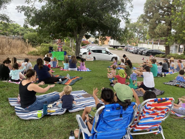 Families watching Sparkles perform an outdoor show beneath a large tree